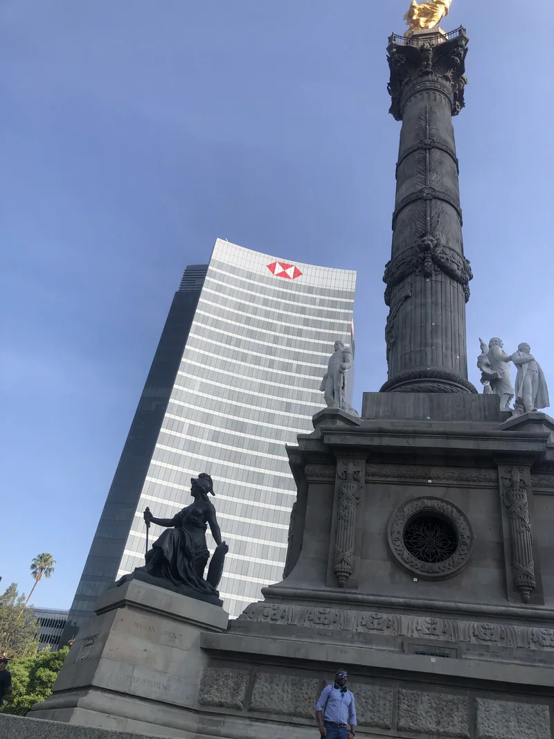 The Ángel de la Independencia column rises above its sculpted base, with a visitor near the bottom and a glass tower behind.