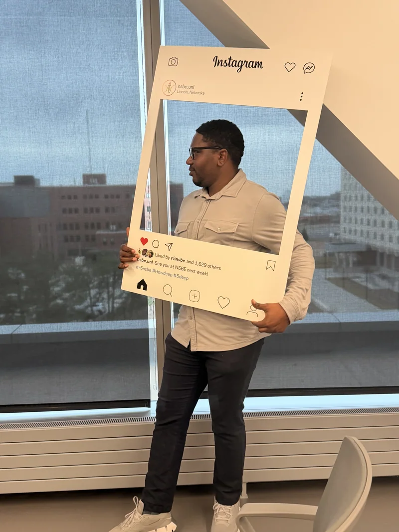 A participant holds an Instagram-style photo frame labeled nsbe.unl and Lincoln, Nebraska beside large campus windows.