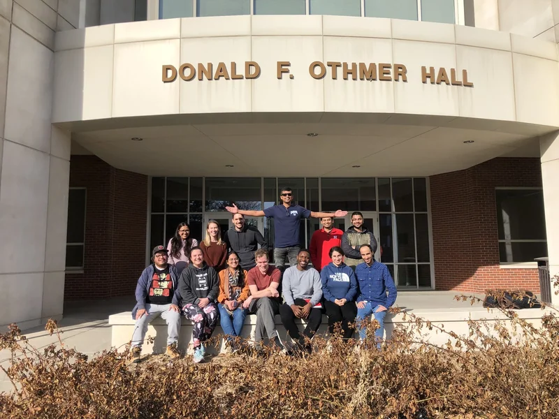 A thirteen-person lab group poses outside the entrance marked Donald F. Othmer Hall.